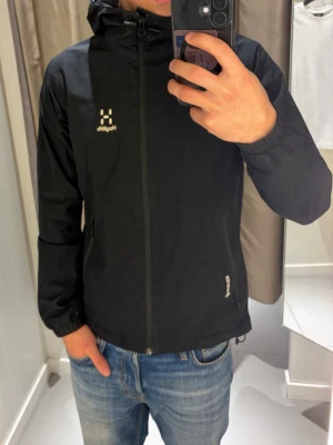 Svart vindjacka från Haglöfs Goretex - Snygg svart vindjacka från Haglöfs Gortex med dragkedja framtill, två sidofickor med dragkedja och justerbar huva. Jackan har elastiska muddar vid ärmsluten och diskret logga på bröstet. Tillverkad i lätt och slitstarkt material, perfekt för blåsiga dagar.