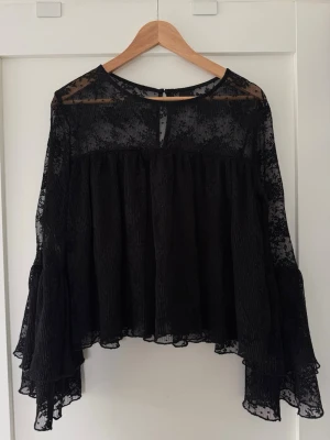 Svart spetsblus från Gina Tricot - Snygg svart blus från Gina Tricot i transparent spetsmaterial. Blusen har långärmade vida ärmar och en lös, flowy passform. Rund halsringning med liten knapp i nacken. Perfekt för dig som gillar trendiga och feminina detaljer.