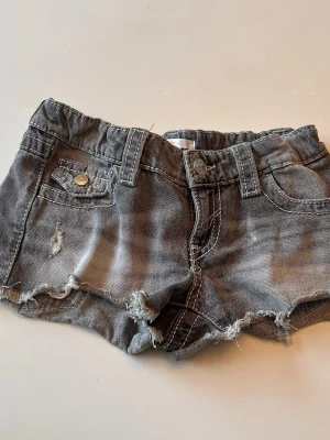 Grå jeansshorts med broderi - Säljer ett par grå jeansshorts med råa kanter och slitningar framtill. Shortsen har dekorativa bakfickor med broderade detaljer och paljetter. Klassisk femficksmodell med knapp och dragkedja. Perfekta för sommaren!