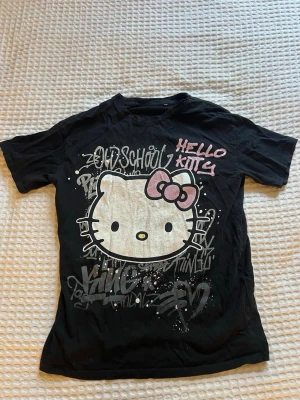Svart Hello Kitty t-shirt - T-shirt med stort Hello Kitty-tryck framtill och graffiti-inspirerad text i vitt och rosa från New Yorker.