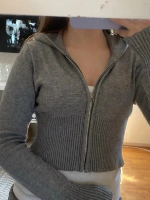 Grå zip hoodie med ribbad midja - Säljer en grå zip hoodie med ribbad midja och långa ärmar. Tröjan har dragkedja framtill och en huva, samt en croppad passform som ger en trendig look. Perfekt att slänga över en t-shirt för en avslappnad stil.