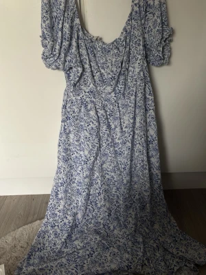 Blåvit långklänning med puffärm - Långklänning i vitt med blått blommönster och puffiga kortärmar. Klänningen har en v-ringad urringning och är tillverkad i ett mjukt bomullsliknande material. Perfekt för sommarens alla tillfällen och ger en romantisk vibe.