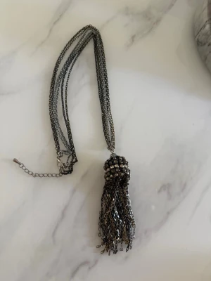 Kedjehalsband med tofs och pärldetaljer - Snyggt halsband i mörk silverfärg med flera kedjor och en stor tofs som hänge. Hänget är dekorerat med små pärlor och ger ett coolt statement-utseende. Perfekt accessoar för att lyfta din outfit med en unik touch.