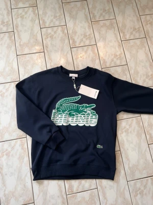 Mörkblå sweatshirt från Lacoste - Snygg mörkblå sweatshirt från Lacoste med stor grön krokodil och vit Lacoste-logga på bröstet. Tröjan har rund halsringning, ribbade muddar och är tillverkad i mjukt bomullsmaterial. Perfekt för en avslappnad och trendig look.