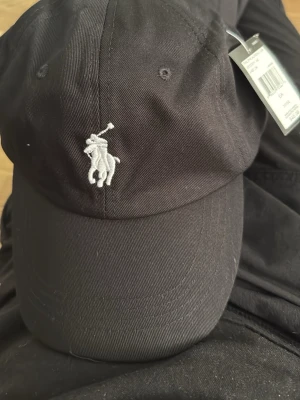 Svart keps från Polo Ralph Lauren - Snygg svart keps från Polo Ralph Lauren med klassisk vit broderad logga framtill och 'POLO' broderat bak. Justerbart spänne i brunt läder och metall. Tillverkad i bomullstyg med böjd skärm och ventilerande öljetter.