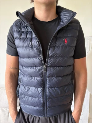 Ralph lauren vest - Snygg mörkblå dunväst från Ralph Lauren med quiltad design och hög krage. Västen har dragkedja framtill, två sidofickor med dragkedja och den klassiska röda loggan broderad på bröstet. Perfekt för lager på lager och streetstyle.