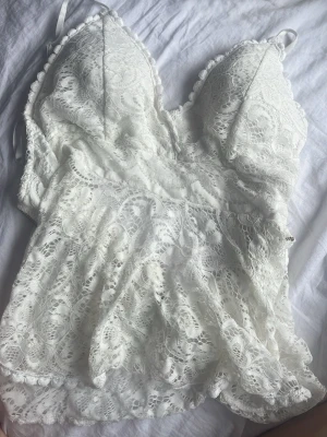 Vit spetslinne med dragkedja lace - Säljer ett vitt spetslinne med tunna axelband och v-ringning. Toppen har fina spetsdetaljer över hela plagget och en dragkedja i ryggen för enkel påklädning. Perfekt för dig som gillar romantisk och feminin stil.