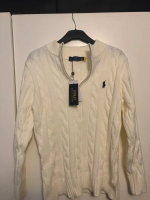 Polo Ralph Lauren Zip Tröja - Kabelstickad vit tröja från Polo Ralph Lauren med halv dragkedja och ribbade muddar. Klassisk logga broderad på bröstet. Tröjan har en hög krage och är tillverkad i mjukt material som passar perfekt till kyligare dagar.
