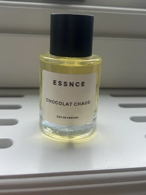 ESSNCE Chocolat Chaud Eau de Parfum - Oanvänd Parfymflaska med guldfärgad vätska från ESSNCE, doften heter Chocolat Chaud. Flaskan är rund med svart lock och vit etikett med svart text. Perfekt för dig som gillar varma, chokladiga doftnoter.