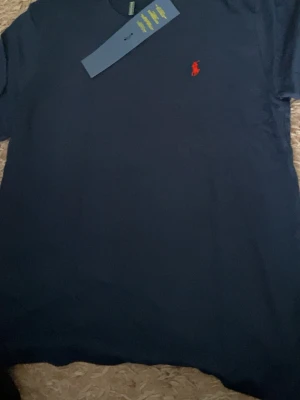Marinblå t-shirt från Ralph Lauren - Snygg marinblå t-shirt från Ralph Lauren med klassisk röd broderad logga på bröstet. T-shirten har rund hals och korta ärmar, perfekt för en stilren och enkel look.