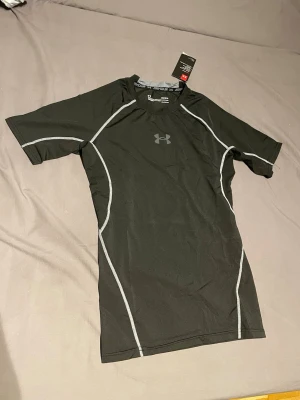 Svart träningströja från Under Armour - Säljer en svart funktions-tshirt från Under Armour med grå kontrastsömmar. Tröjan har korta ärmar och är tillverkad i ett stretchigt syntetmaterial som andas, perfekt för intensiva träningspass. Under Armour-logga på bröstet och nacken.