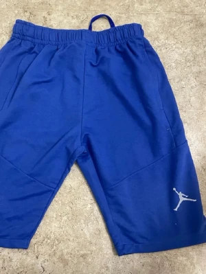 Blå Jordan shorts med logga - Säljer ett par blå shorts från Jordan med den klassiska Jumpman-loggan i vitt på ena benet. Shortsen har elastisk midja och är tillverkade i mjukt material som passar perfekt för sportiga aktiviteter.