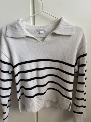 Vit stickad tröja Gina Tricot - Vit stickad tröja från Gina Tricot med krage och v-ringning. Ena ärmen har svarta horisontella ränder som ger en cool kontrast. Perfekt till jeans eller kjol för en avslappnad men trendig look.