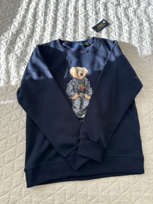 Mörkblå sweatshirt Polo Ralph Lauren - Mörkblå sweatshirt från Polo Ralph Lauren med ikoniska Polo Bear-trycket framtill. Rund halsringning, ribbade muddar vid ärmslut och nederkant. Tillverkad i mjukt bomullsmaterial för en skön känsla. Perfekt för en avslappnad och trendig stil.