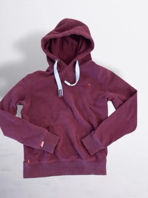 Vinröd Superdry Hoodie - Säljer en vinröd hoodie från Superdry med klassisk känguruficka och kontrasterande ljusa dragsnören. Hoodien har ribbade muddar vid ärmslut och nederkant samt en liten logga på bröstet. Perfekt för en avslappnad och snygg stil.