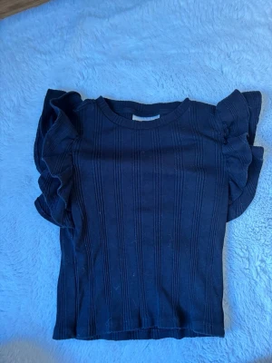 Svart ribbad topp från Gina Tricot - Snygg svart ribbad topp från Gina Tricot med volangdetaljer på axlarna. Toppen har rund halsringning och är tillverkad i ett stretchigt material som sitter bekvämt. Perfekt för dig som vill ha en trendig och stilren look.