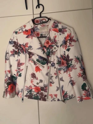 Vit blommig skinnjacka från My Wear - Snygg vit skinnjacka från My Wear med färgglatt blommönster i rött, lila och grönt. Jackan har klassisk bikerstil med sned dragkedja, två framfickor med dragkedjor och dekorativa knappar. Perfekt för dig som vill sticka ut med en unik look.