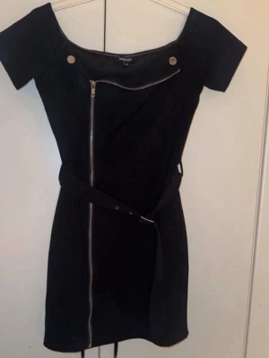 Svart kort klänning med dragkedja - Snygg svart kort klänning från Boohoo med asymmetrisk dragkedja framtill, dekorativa knappar vid axlarna och bälte i midjan. Klänningen har korta ärmar och en figurnära passform som ger en cool look.
