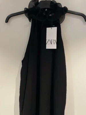 Svart ärmlös top från Zara - Stilren svart top från Zara med hög hals och volangdetaljer runt kragen. Topen  är ärmlös och har en rak, elegant siluett som ger en modern look. Perfekt för dig som gillar minimalistisk design med en twist.