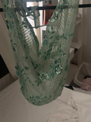 Mintgrön paljettprydd halterneck topp - Säljer en mintgrön halterneck topp med blommiga paljettdetaljer och transparent mesh. Toppen har knyt i nacken och är dekorerad med glittriga broderier och paljetter som ger en unik look. 