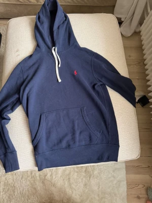 Mörkblå hoodie från Ralph Lauren - Mörkblå hoodie från Ralph Lauren med vit dragsko i huvan och klassisk känguruficka framtill. Liten röd logga broderad på bröstet. Tillverkad i mjuk bomull för en skön och avslappnad look. Hyfsat bra skick, säljs pga för liten. Bara höra av sig vid frågor 
