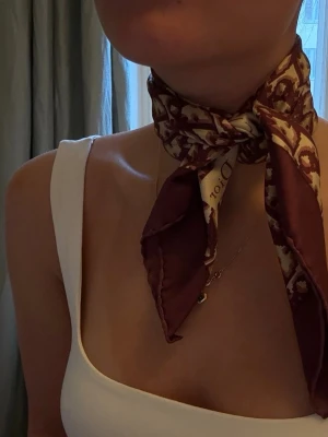 Sidenscarf Dior - Säljer en lyxig bordeauxfärgad sidenscarf från Christian Dior med ikoniskt monogrammönster och bred kant. Scarfen är kvadratisk och har en elegant vintagekänsla, perfekt att knyta runt halsen för en trendig look.