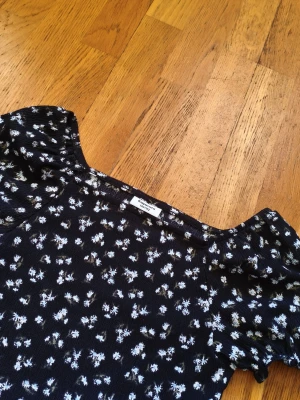Svart blommig topp - Supersöt svart topp från Kappahl med vita blommor och korta puffärmar. Den har en fyrkantig halsringning och volang nertill som ger en extra touch. Perfekt för dig som gillar en feminin och trendig stil. Materialet känns lätt och bekvämt.