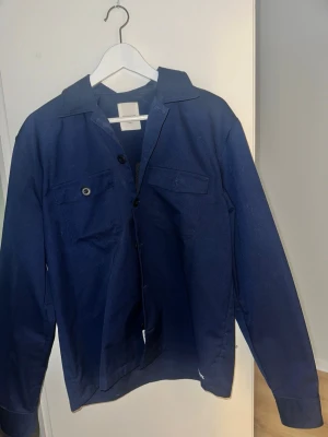 Mörkblå overshirt med fickor - Stilren mörkblå overshirt med klassisk krage och två bröstfickor med lock. Långa ärmar och knäppning framtill. Perfekt att bära öppen över en t-shirt eller stängd som en lätt jacka. Wood Wood är märket. 