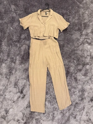 Beige set med kortärmad topp och byxor - Snyggt beige set med en kortärmad, knäppt topp och matchande höga byxor i mjukt, luftigt tyg. Toppen har krage och korta ärmar, byxorna har avslappnad passform och resår i midjan. Perfekt för en chill och trendig look.