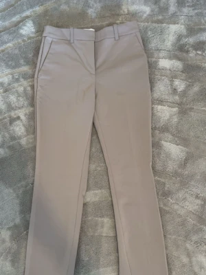 Beige kostymbyxor med raka ben - Snygga beige kostymbyxor med raka ben och pressveck. Byxorna har hällor för skärp och sidofickor. Perfekta för en clean och stilren look. Materialet känns mjukt och har en lätt glans. Märke och storlek syns inte på bilderna.
