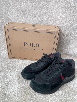 Svarta Polo Ralph Lauren sneakers - Snygga svarta sneakers från Polo Ralph Lauren med röd broderad logga på sidan. Skorna har en mix av mocka och textilmaterial, klassisk låg siluett och svarta skosnören. Perfekta för en stilren och sportig look.
