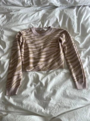 Beige zebramönstrad stickad tröja - Säljer en croppad stickad tröja i beige och ljusrosa med zebramönster. Tröjan har rund halsringning, långa ärmar och ribbade muddar vid ärmslut och nederkant. Perfekt för dig som gillar trendiga mönster och mjuka material.