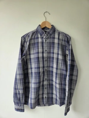 Blårutig skjorta från Tiger of Sweden - Snygg blårutig skjorta från Tiger of Sweden med klassisk button-down krage och långa ärmar. Skjortan har knappar framtill och är tillverkad i mjuk bomull som känns skön mot huden. Perfekt för dig som gillar stilrena och tidlösa plagg.