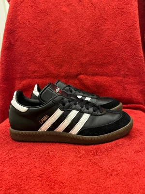 Adidas Samba svarta sneakers EUR 38 - Klassiska Adidas Samba sneakers i svart läder med vita detaljer och de ikoniska tre ränderna på sidorna. Skorna har gummisula, svart mocka vid tån och vit hälflik. Perfekta för en stilren och sportig look.