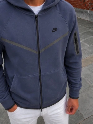 Blå zip hoodie från Nike - Snygg blå hoodie från Nike med dragkedja framtill och svarta detaljer. Har huva, fickor på magen och en extra ficka med dragkedja på ärmen. Nike-logga på bröstet och diskreta reflexband över bröstet. Perfekt för en sportig och avslappnad stil.