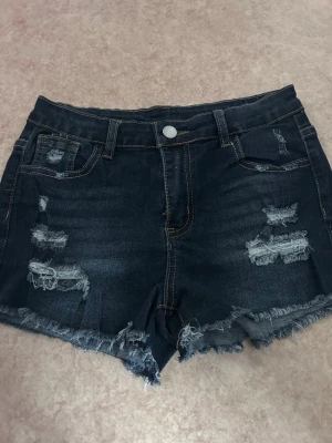 Mörkblå slitna jeansshorts - Snygga mörkblå jeansshorts med slitna detaljer och fransig kant. Klassisk femficksmodell med knapp och dragkedja. Perfekta för sommaren och ger en avslappnad streetstyle-look.