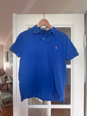 Blå pikétröja från Ralph Lauren - Klassisk blå pikétröja från Ralph Lauren med orange broderad logga på bröstet. Tröjan har korta ärmar, krage och knappar vid halsen. Perfekt till jeans eller shorts för en clean och stilren look.