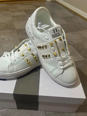 Valentino Rockstud vita sneakers - Snygga vita sneakers från Valentino med guldiga nitar på sidorna och breda tygband. Skorna har en klassisk låg siluett och är tillverkade i läder med detaljer i textil. Perfekta för dig som vill sticka ut med en lyxig och edgy stil.
