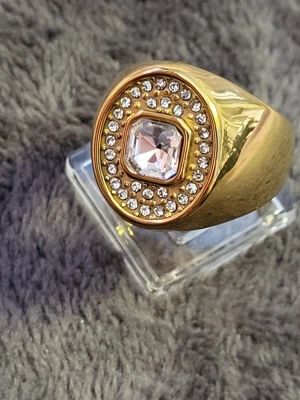 Guldfärgad klackring med ljusrosa glittrande stenar - Snygg guldfärgad kraftig klackring med en stor fyrkantig ljusrosa sten i mitten, omgiven av två rader små gnistrande stenar. Ringen har en bred och blank design som verkligen drar blickarna till sig. Perfekt för dig som gillar statement-smycken och vill ha något som sticker ut.