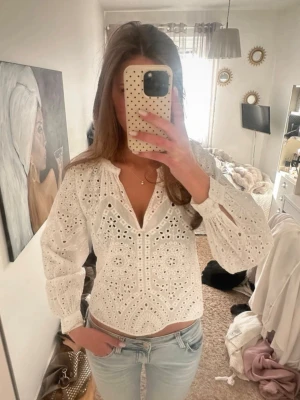 Vit broderad blus med v-ringning - Säljer en vit blus med v-ringning och broderade hålmönster. Blusen har långa ärmar med resår vid ärmslut och en avslappnad passform. Tillverkad i ett lätt bomullstyg som ger en luftig känsla och romantisk look. Passa mig som bruka ha xs-s