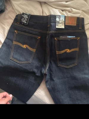 Nudie Jeans Dry Dirt Organic - Säljer ett par mörkblå jeans från Nudie Jeans i modellen Dry Dirt Organic. Jeansen är tillverkade av ekologisk bomull och har klassiska kontrastsömmar samt Nudie Jeans ikoniska broderi på bakfickorna. Designade i Sverige och tillverkade i Italien.