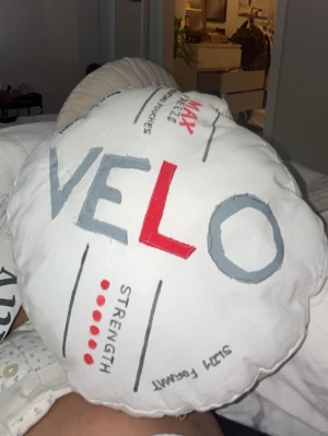Vit rund kudde med VELO-tryck - En rund, vit kudde med tryck i grått, rött och svart där det står VELO och olika grafiska detaljer. Kudden är mjuk och har en lekfull design som påminner om en snusdosas lock. Gjorde i slöjden i 9 an 