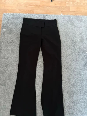 Svarta kostymbyxor från Gina Tricot - Snygga svarta kostymbyxor från Gina Tricot med utsvängda ben. Byxorna har en klassisk knappstängning och är tillverkade i ett mjukt, stretchigt material som sitter bekvämt. Perfekta för en stilren look. Några få defekter men inga som syns jätte tydligt. Skriv om mer bilder!