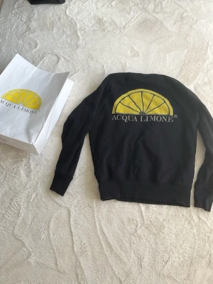  Acqua Limone sweatshirt  - Säljer en svart sweatshirt från Acqua Limone med stort gult citrontryck och logga på ryggen samt texten 'Acqua Limone classic' på bröstet. Tröjan har rund halsringning, ribbade muddar och är tillverkad i mjukt bomullsmaterial.
