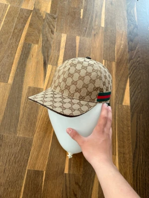 Gucci beige keps  - Säljer en beige keps från Gucci med det klassiska GG-monogrammönstret över hela kepsen. På sidan finns en grön och röd rand som signaturdetalj. Kepsen har böjd skärm och justerbar rem bak. Materialet är canvasliknande tyg.