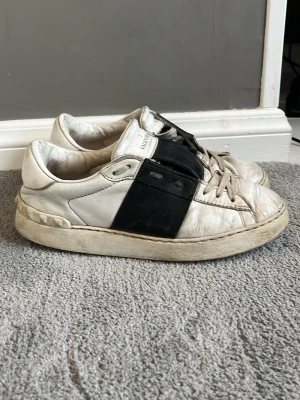 Vit/svart sneaker från Valentino - Snygga sneakers från Valentino i vitt läder med en bred svart läderdetalj över sidan och svarta snören. Klassisk låg modell med chunky sula och diskret logga på plösen. Perfekta för dig som gillar stilrena och exklusiva sneakers.