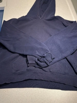 Mörkblå hoodie med magficka - En klassisk mörkblå hoodie med huva och stor magficka framtill. Hoodien har ribbade muddar vid ärmslut och nederkant. Perfekt för en avslappnad och stilren look. Materialet känns mjukt och bekvämt, troligtvis bomullsblandning.