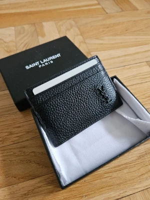 Svart Y S L kortfodral i läder - Snyggt svart kortfodral från Saint Laurent i exklusivt präglat läder. Fodralet har flera kortfack och den ikoniska loggan i svart framtill. Perfekt för dig som vill ha en stilren och lyxig accessoar i fickan.