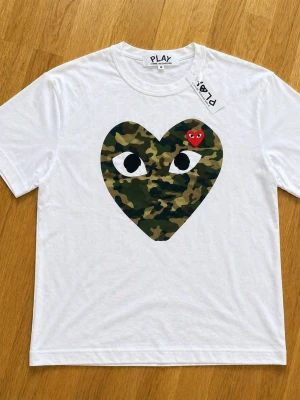 CDG t shirt  - Vit t-shirt från Comme des Garçons PLAY med ett stort hjärta i camouflage och ögon på bröstet samt ett litet rött hjärta. Klassisk passform och rund halsringning. Perfekt statement-plagg för dig som gillar streetwear.