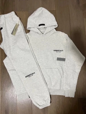ESSENTIALS FEAR OF GOD ’LIGHT OATMEAL’ (SS22) - Tracksuit från Essentials Fear of God i ljusgrått. Setet består av en hoodie med skön 80/20 Poly, Ull blend och matchande mjukisbyxor med resår i midja och benslut. Båda plaggen har svart Essentials-logga tryckt framtill. Perfekt för chill dagar hemma. Finns inne just nu📲